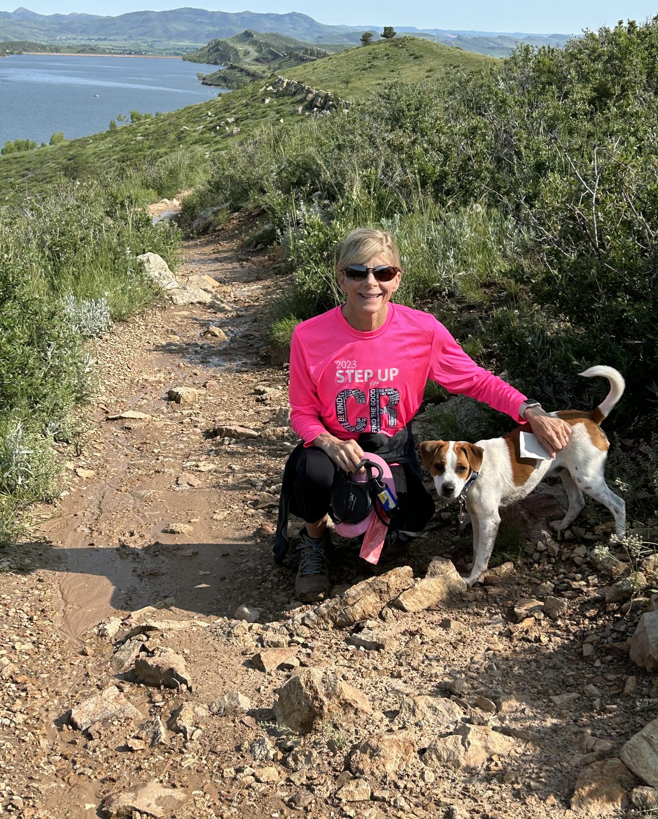 Nina Caron crouching beside her dog on a rocky trail above a lake, wearing her Step Up for Colleen shirt